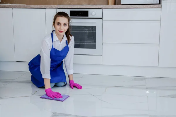Gyönyörű fiatal lány mossa a padlót a kezét rózsaszín kesztyűben. A lány belenéz a keretbe. A háttérben egy gyönyörű fehér modern konyha — Stock Fotó
