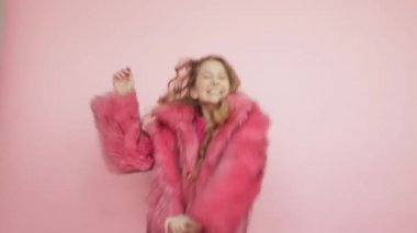 Uzun saçlı genç bir kız pembe arka planda pembe bir ceketle dans ediyor..