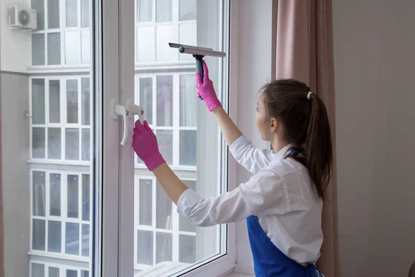 Hermosa chica joven lava una ventana. Fuera de la ventana hay edificios de varios pisos . — Foto de Stock