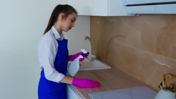 Uma menina morena com uma camisa branca e um avental azul está limpando a cozinha com um detergente . — Vídeo de Stock