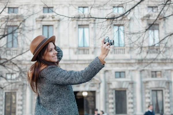 Молодая брюнетка фотографирует, держа шляпу рукой. На фоне серого старого здания . — стоковое фото