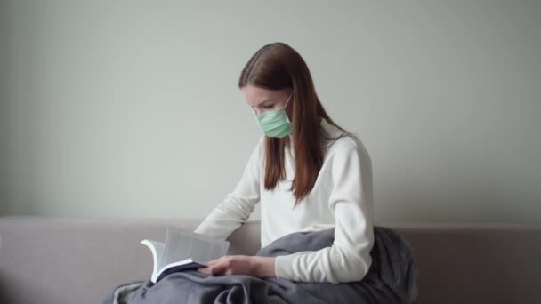 Egy fiatal lány, aki orvosi maszkban ül a kanapén könyvet olvas.. — Stock videók