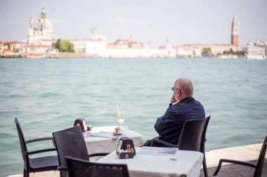 Bir adam restoranda kokteyl içer ve deniz koyuna bakar..