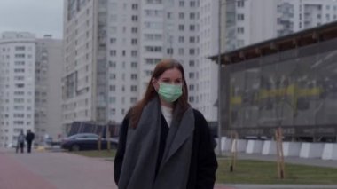 Yüzünde tıbbi maske olan uzun saçlı güzel bir genç kız şehirde dolaşıyor. Gri gökdelenlerin arka planına karşı. Orvi, koronovirüs, grip, akut solunum hastalığı..