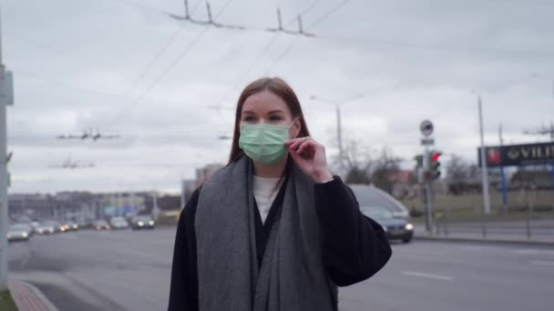 Beautiful young girl with long hair straightens a medical green mask against the background of passing cars. Orvi, coronovirus, flu, acute respiratory disease. — Stock Video