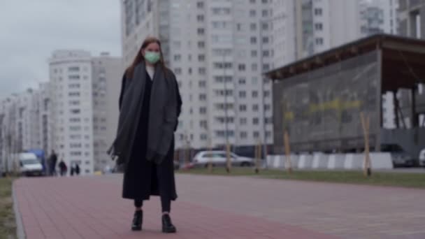 Una hermosa joven con el pelo largo con una máscara médica en la cara camina por la ciudad. En pleno crecimiento. En el fondo de edificios grises de gran altura. Orvi, coronovirus, gripe, aguda — Vídeo de stock