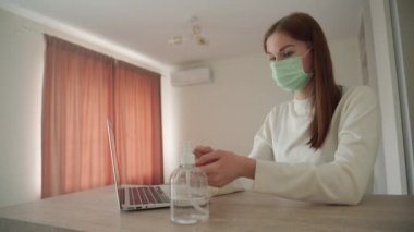 Güzel genç kız evde çalışıyor ve ellerini dizüstü bilgisayarın yanında bir antiseptikle işliyor. Orvi, koronovirüs, grip, akut solunum hastalığı..