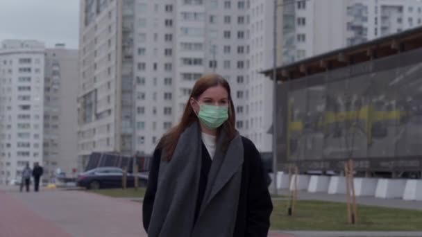 Ein schönes junges Mädchen mit langen Haaren und einer medizinischen Maske auf dem Gesicht geht durch die Stadt. Vor dem Hintergrund grauer Hochhäuser. Orvi, Coronovirus, Grippe, akute Atemwegserkrankungen. — Stockvideo