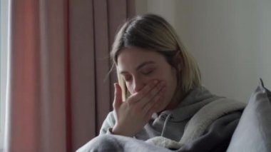Sarışın kız gri bir kanepede oturuyor, viral hastalıklardan hasta ve avucunda öksürüyor. Orvi semptomu, koronovirüs, grip, akut solunum hastalığı, alerji, sinüzit..