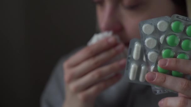 Уставшая больная девушка показывает лекарства на камере крупным планом, держит в руке много лекарств и чихает. Зеленые, белые большие таблетки . — стоковое видео