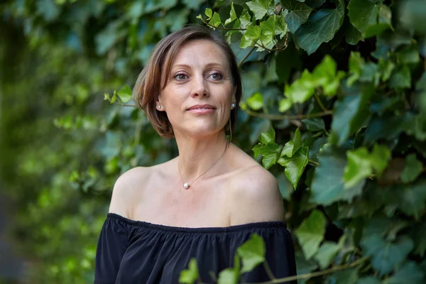 Atractiva mujer en un vestido negro con hombros desnudos y un collar de perlas posando cerca de una pared verde brillante de plantas . —  Fotos de Stock