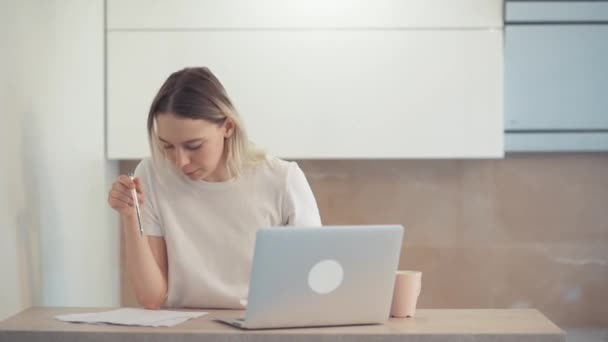 Egy fiatal lány koncentrál a laptopjára, és összehasonlítja az információt azzal, ami egy darab papírra van írva. Fényes tágas szoba. — Stock videók