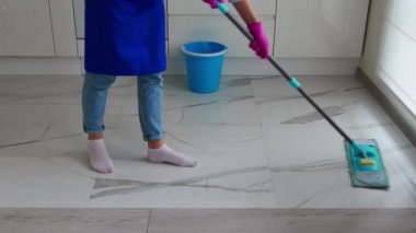 Mavi önlüklü ve mavi kotlu bir kızın mavi paspasla temizlik yaptığı görüntüler. Yakınlarda bir kova var..