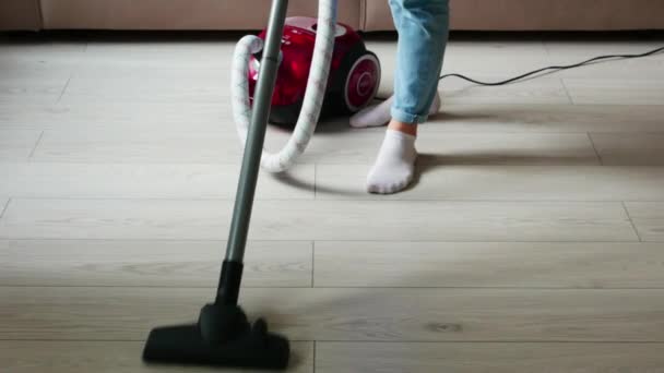 Gambar seorang gadis dengan celana jeans biru yang sedang membersihkan, menyedot debu di lantai kayu dengan vacuum cleaner merah. — Stok Video