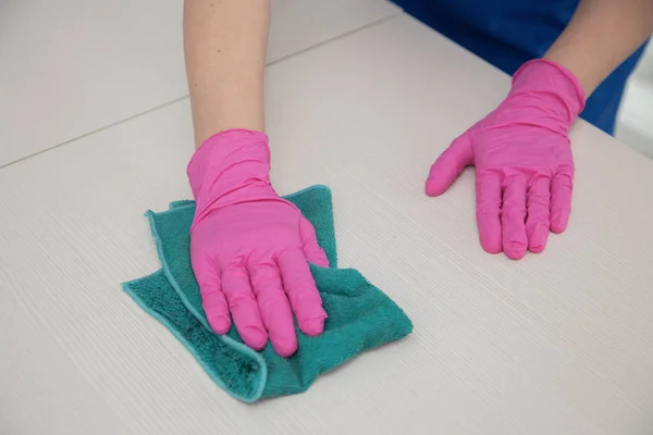 Een hand in een roze handschoen veegt het witte oppervlak van de tafel af met een turquoise poetsdoek. — Stockfoto