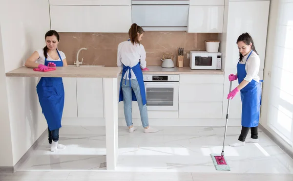 Limpeza de casa profissional. Três meninas bonitas estão limpando a cozinha. A menina lava o chão . — Fotografia de Stock