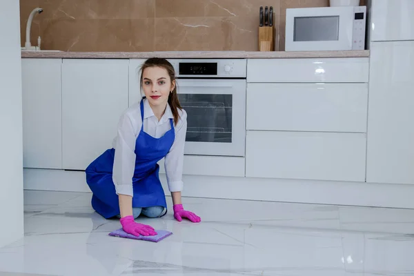 Gyönyörű fiatal lány mossa a padlót a kezét rózsaszín kesztyűben. A háttérben egy gyönyörű fehér modern konyha. — Stock Fotó