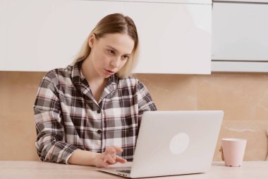 Genç ve güzel bir kız elinde kahve fincanıyla dizüstü bilgisayarın önünde oturuyor..