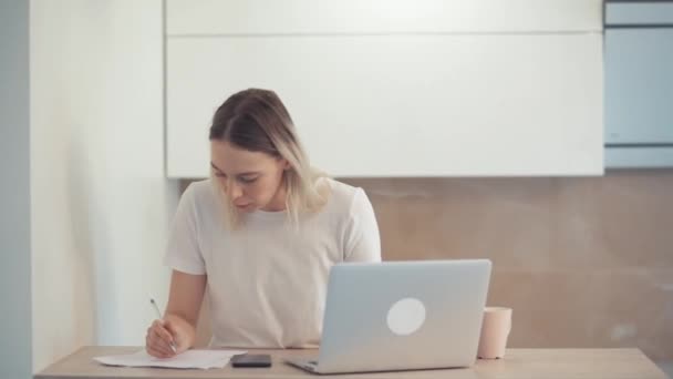Una joven se concentra en un ordenador portátil y describe una hoja de papel blanco. Lea en la calculadora en el teléfono . — Vídeos de Stock