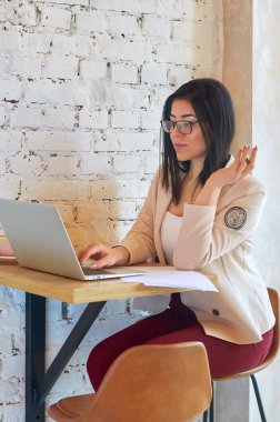 Gözlüklü kız, beyaz dizüstü bilgisayarlı, modaya uygun bir kafede çalışıyor. Açık renk ceketli ve kırmızı pantolonlu kız..