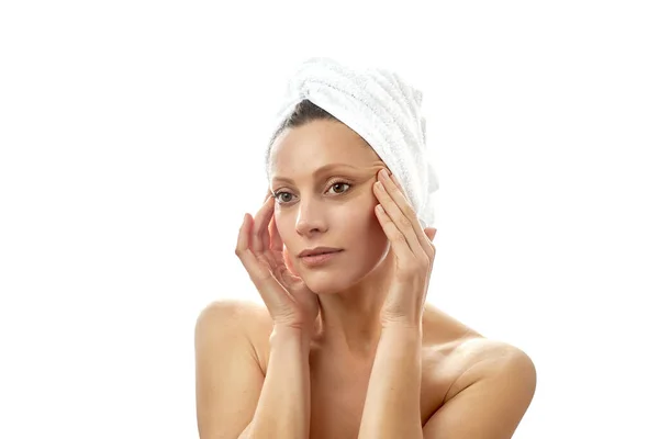 Mulher bonita com uma toalha branca na cabeça e com pele limpa e perfeita faz massagem facial em casa. Massageie a pele ao redor dos olhos. Fundo branco isolado . — Fotografia de Stock
