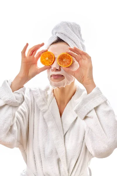Seorang wanita cantik dalam mantel putih dan handuk menempatkan irisan jeruk segar ke wajahnya dan berpaling. Latar belakang putih terisolasi. — Stok Foto