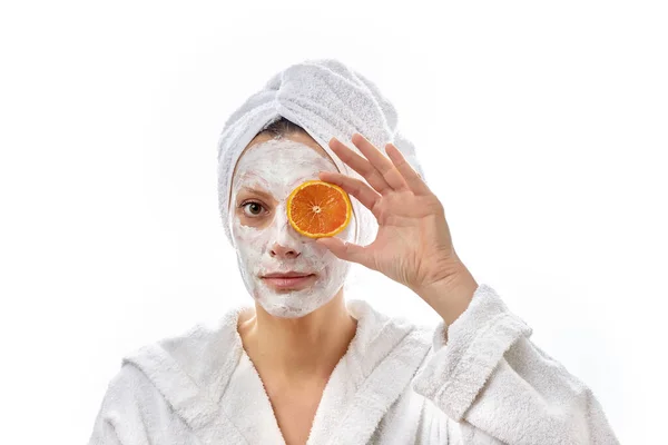 Piękny portret dziewczyny z białym ręcznikiem na głowie i w białym płaszczu trzymającej pomarańczowe plasterki na twarzy i patrzącej w kamerę. Izolacja na białym tle. — Zdjęcie stockowe