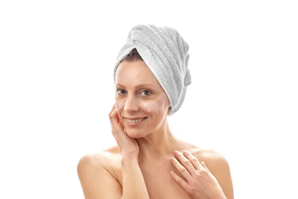 Krásná žena s bílým ručníkem na hlavě dělá omlazení obličeje s páskou přes oko a usmívá se do kamery. Bílé izolované pozadí. — Stock fotografie