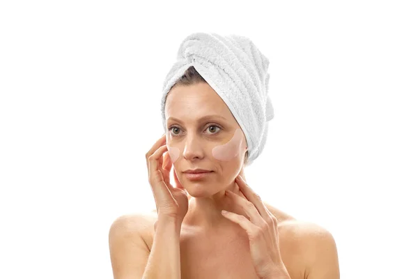 Krásná žena s bílým ručníkem na hlavě dělá omlazení obličeje páskou přes oko. Bílé izolované pozadí. — Stock fotografie
