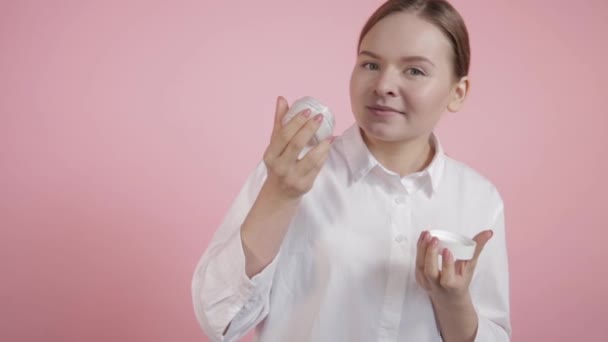 Chica huele una crema blanca en un frasco de vidrio sobre un fondo rosa — Vídeos de Stock