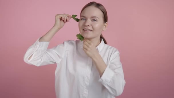 Hermosa joven morena en una camisa blanca sobre un fondo rosa sin maquillaje con hierbas en las manos . — Vídeos de Stock