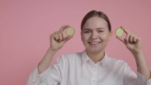 Una joven sin maquillaje tiene en sus manos dos rodajas de pepino cerca de sus ojos . — Vídeos de Stock