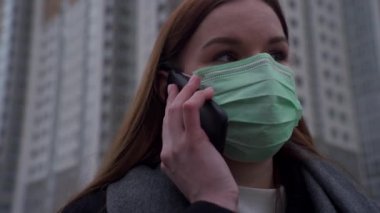 Yakın plan, uzun saçlı, yüzünde tıbbi maske olan güzel bir kız. Yüksek binaların arka planında telefonla konuşuyor..