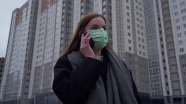 Uzun saçlı, yüzünde tıbbi bir maske olan genç bir kız yüksek binaların arka planında telefonla konuşuyor..