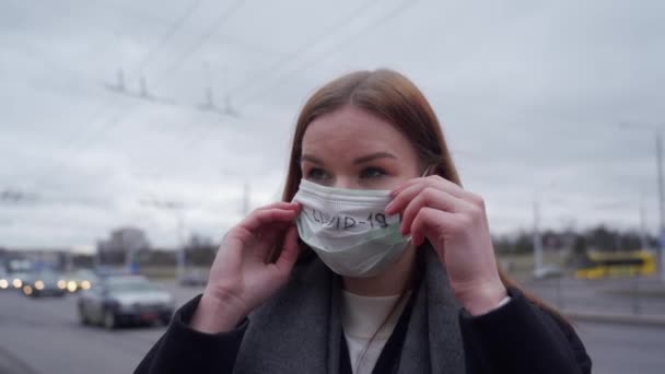 Mooi meisje met lang haar rechtzet een medisch groen masker met de inscriptie covid-19 op de achtergrond van passerende auto 's. — Stockvideo