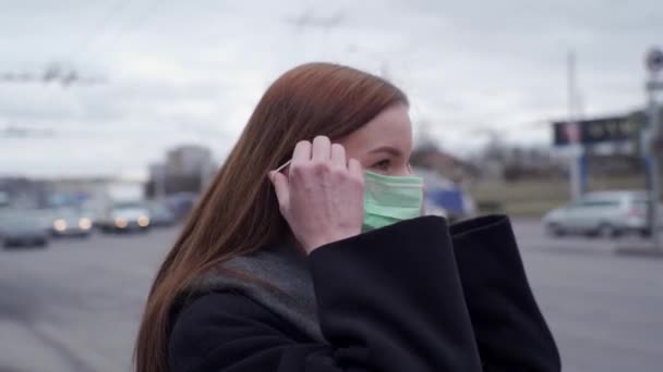 Vacker ung flicka med långt hår bär en medicinsk grön mask mot bakgrund av passerande bilar. Sidovy. — Stockvideo