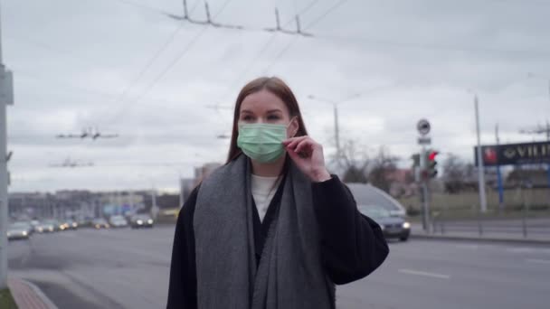 Gyönyörű fiatal lány hosszú hajjal egyenesbe hozza az orvosi zöld maszkot az elhaladó autók hátterében.. — Stock videók