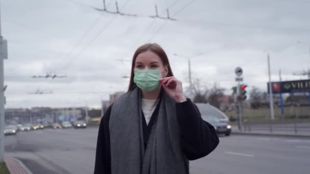 Hermosa joven con el pelo largo endereza una máscara verde médica en el fondo de los coches que pasan . — Vídeo de stock