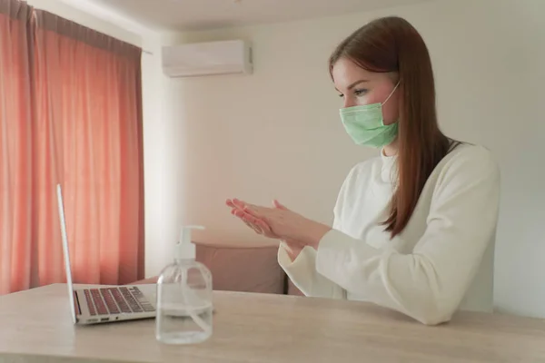 A beautiful girl works from home in a medical mask and rubs an antiseptic in her hands. — Stock Photo, Image