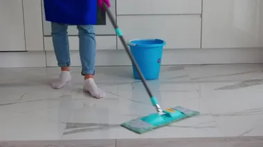 Mavi önlüklü ve mavi kotlu bir kızın mavi paspasla temizlik yaptığı görüntüler. Yakınlarda bir kova var..