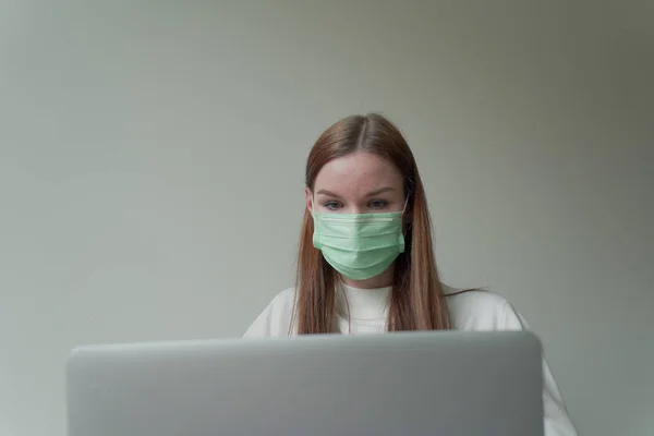 Joven chica hermosa en una máscara médica sostiene un ordenador portátil en sus manos. Trabajo desde casa durante la cuarentena . —  Fotos de Stock