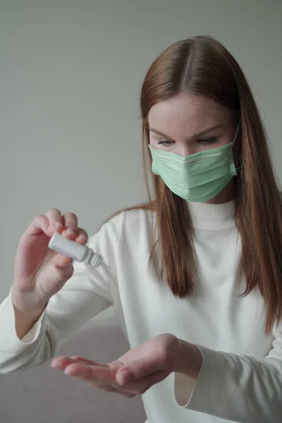 Una joven con una máscara médica se sienta en un sofá y usa un pequeño frasco de bolsillo de antiséptico. Desinfección de manos. Foto vertical . —  Fotos de Stock