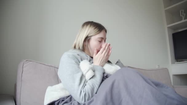 Het blonde meisje zit op een grijze bank gewikkeld in een deken is ziek, hoest in de palm van haar hand. — Stockvideo