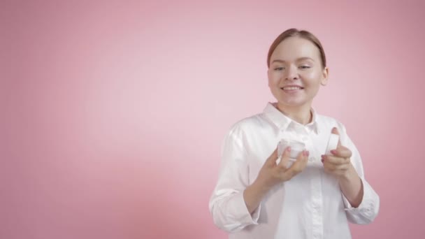 Una joven con una camisa blanca sobre un fondo rosa abre un frasco de crema blanca — Vídeos de Stock