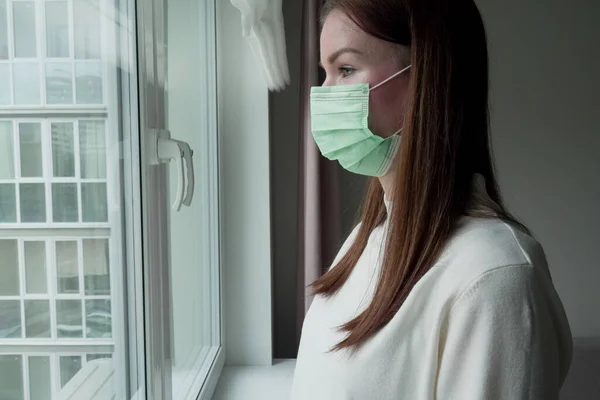 Una joven con una máscara médica se ve tristemente por la ventana. La chica en cuarentena extraña a su familia y amigos . —  Fotos de Stock