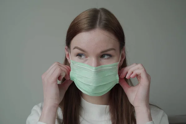 Primer plano de una hermosa joven con el pelo largo y rojo en una máscara médica. La chica mira hacia la ventana. Orvi, coronovirus, gripe, infecciones respiratorias agudas . —  Fotos de Stock