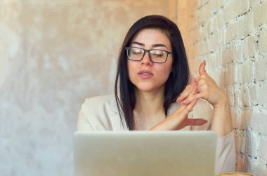 Gözlüklü esmer bir öğrenci, beyaz tuğla bir duvarın arka planında dizüstü bilgisayarla çevrimiçi öğrenim görüyor..