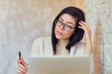 Gözlüklü esmer bir öğrenci, beyaz tuğla bir duvarın arka planında dizüstü bilgisayarla çevrimiçi öğrenim görüyor..