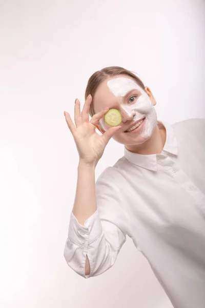 Schöne junge Mädchen mit Feuchtigkeitscreme auf dem Gesicht. Eine Gurkenscheibe halten. — Stockfoto