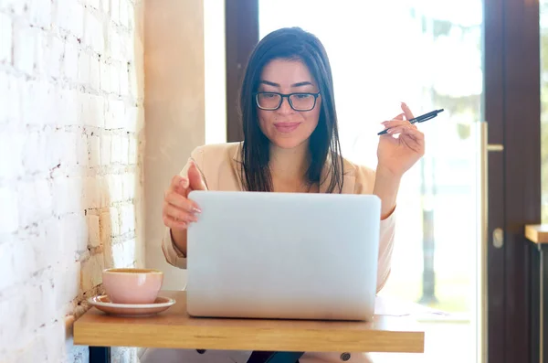 Güzel esmer kadın bir kafede laptopun başında çalışıyor ve bir fincan taze aromatik kahvenin yanında..